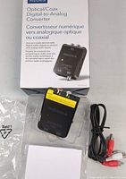 Photo of Insignia Optical/Coax Digital-to-Analog Converter showing front panel and included RCA cable in plastic packaging with box behind.