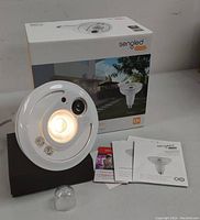 Front view of Sengled Snap LED lamp showing LED light, HD camera lens, and sensor components with box and manuals in background.