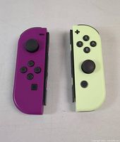 Front view of two Nintendo Switch Joy-Con controllers side-by-side, purple left and light green right, showing buttons and analog sticks.