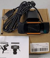 Front view of the black compact webcam with orange trim on a cardboard box alongside the USB cable coiled beside it. A printed instruction manual is partially visible.