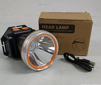 Photo showing the head lamp, USB cable, and box. The head lamp features a large circular reflector with an orange rim and adjustable strap.