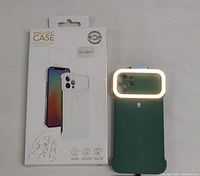 Photo showing one ringlight phone case illuminated, positioned upright beside its retail box. The ringlight is turned on, glowing white around the camera area of the case.