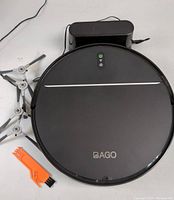 Top view of the BAGO robot vacuum cleaner showing power and WiFi indicators, charging dock, two replacement side brushes, and orange cleaning tool on a white surface.