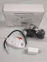 White Refoss Smart Wi-Fi Garage Door Opener device with wiring and power adapter laid out in front of original retail box showing product name and usage.