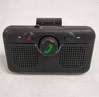 Front view of the black stereo car speakerphone showing speaker grille and control buttons including a large central call button, mute, track controls, and call answer/end buttons