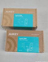 Two brown cardboard boxes each labeled 'Swift 20W USB-C PD Charger' from Aukey with blue sticker showing product outline and branding, indicating sealed new condition.