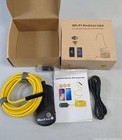 Open box showing WiFi endoscope unit with coiled yellow flexible cable, USB charging cable, and instruction manual next to original cardboard box.