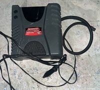 Photo of first Motomaster air compressor with digital display and attached cables and clamps lying on floor, black and gray plastic body.