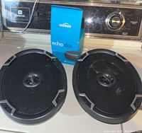 Two oval black speakers with metal grills and central tweeter alongside Amazon Echo Dot box on a surface.