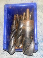 Top view of a blue container holding several very large metal drill bits with visible shanks and spiraled cutting edges, showing some surface rust and dirt.