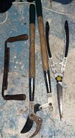 Three garden cutting tools on a blue floor including a brace and bit hand drill, long-handled hedge/shears, and pruning shear, all with wooden handles and showing rust.
