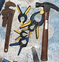 Photo of a claw hammer with a wooden handle, rusty pipe wrench, and four plastic spring clamps with yellow and blue grips on a blue surface.