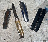 Four pocket utility knives laid out on scratched metal surface, showing handles and blades.