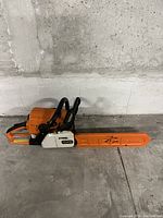 Right side profile of orange Stihl chainsaw with protective bar cover on concrete floor against concrete wall