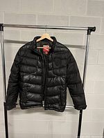 Front view of black men's medium puffer coat hanging on a rack against a concrete wall.