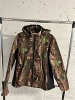 Front view of a camouflage patterned coat with hood hanging on a rack against a white wall.