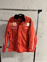 Front view of red jacket on rack showing FILA and Ferrari patches, sponsor logos and zip pockets