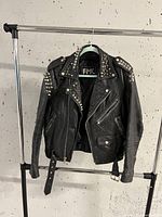 Front view of black leather motorcycle jacket with silver studs on collar, shoulders, and lapels, front asymmetrical zipper, pockets, and waist belt visible.