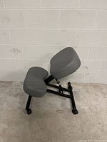 Photo of gray ergonomic kneeling chair with padded seat and knee rests, adjustable backrest, black metal frame, and caster wheels.