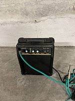 Front view of small black guitar amplifier with control knobs and input jack with cable connected.