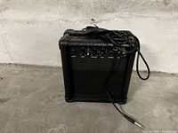 Black guitar amplifier with control panel, knobs, and attached cable against a concrete floor and wall.