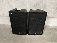 Front view showing two trapezoidal black speaker cabinets with hexagonal metal grills and small logo on each grill.