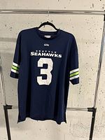 Front view of navy blue Seattle Seahawks jersey with number 3 and team name on front. Hanging on black hanger against white wall.