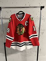 Front view of the Chicago Blackhawks Jonathan Toews hockey jersey with team logo, captain 'C', and number '19'. Hanging on rack against a concrete wall.