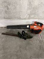 Photo shows two garden tools on concrete floor against a wall: An orange and black Echo leaf blower and a green Greenworks hedge trimmer with metal blades, both without cords.