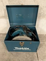 Blue Makita circular saw inside an open metal carrying case showing the saw blade and cord.