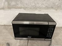 Front view of black NuWave Chef microwave oven with stainless steel front edges, digital keypad on right, glass door with black frame, sitting on concrete floor near wall.
