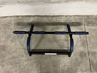Top view of black metal pull up bar with blue and black foam grips and elbow rests on a concrete floor.