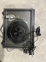 Black record player turntable viewed from above on a concrete floor, shows transparent dust cover, tonearm, cables, and power cord