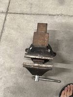 Photo of a metal bench vise with rust and signs of wear, showing the top view including the screw handle and mounting holes.