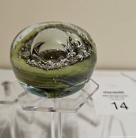 Front angle view showing the smooth glass dome encasing a metallic silver orb with textured bubbles and olive green background inside the paperweight on an acrylic stand.