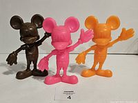 Three vintage Mickey Mouse figures in brown, pink, and orange plastic, posed waving, with noted markings and colors visible.