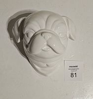Front-facing view of glossy white ceramic pug head wall art mounted on wall with auction lot number 81.