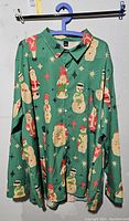 Front view of the green XXXL shirt with various Christmas blow mold motifs including Santa, snowmen, holly,and Christmas trees on a green background hung on a blue hanger.