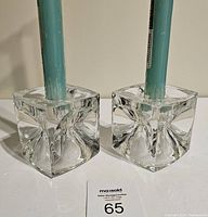 Close-up view of two clear glass cube candle holders, showing their textured internal design that refracts light. Both hold a blue-green tapered candle.