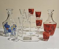 Full view of both decanter sets and accompanying glasses displayed on a white surface, showing floral and red/black scenic designs.
