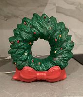Front view of the vintage ceramic wreath lamp in green with colored decorative lights and a bright red bow base, placed on a grey counter.