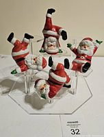 Four vintage ceramic Santa Claus figurines in various exercise poses placed on clear acrylic stands, with lot number visible