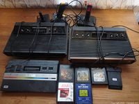 Wide view showing two woodgrain consoles, one Jr console, two joysticks and six cartridges on table