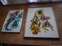 Two framed needlework artworks placed side by side on wood surface. One has a parrot image, the other a ladder with plants.