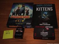 Lot photo showing Pandemic board game box, Imploding Kittens expansion box, Exploding Kittens card game box, and I've Never Bar Cards deck on a wooden surface with a lot number sticky note.