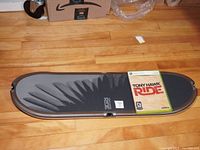 Photo of black and gray skateboard accessory for Nintendo Wii lying on wooden floor with Xbox 360 Tony Hawk Ride game case on top.