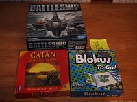 Photo showing three board games boxed: Battleship, Settlers of Catan Portable Edition, Blokus To Go on a floor surface.