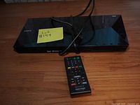 Sony BDP-S5100 Blu-ray player with remote control and power cable on wooden surface, label with lot number visible.