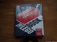 Front cover of 'The Lego Book' coffee table book sealed in shrink-wrap with a label mentioning an exclusive Lego brick and print included.