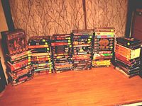 Five stacks of VHS tapes displayed on a table. Tapes feature various movie and concert titles, with colorful covers.